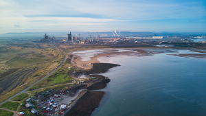 Aerial photo of beach leading to power plant with hills in the background.
